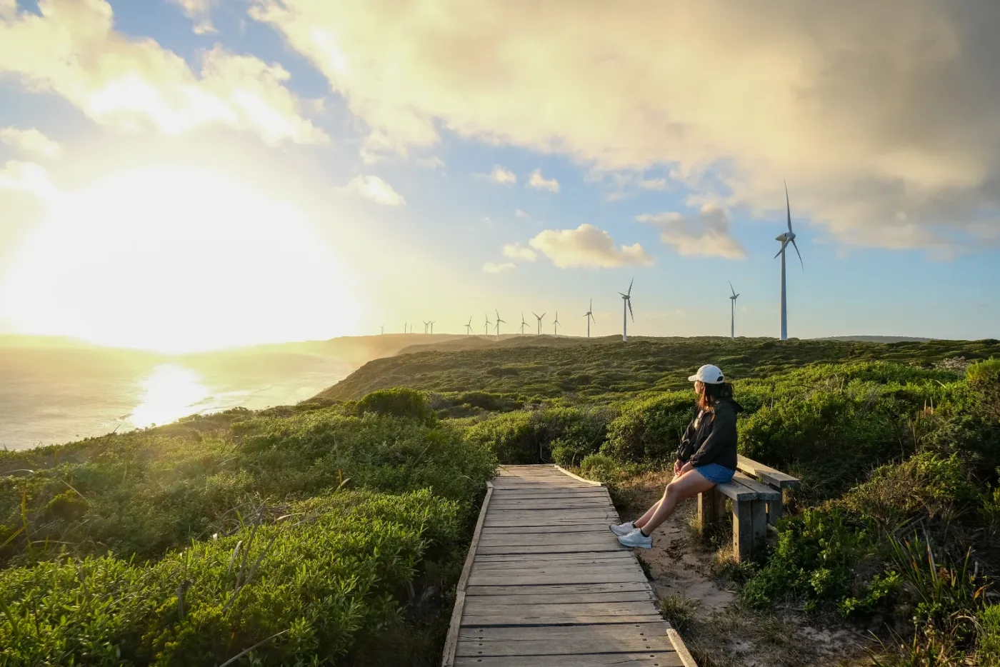 A traveller resting on a boardwalk above the coast near Albany at sunset, with wind turbines on the hillside beyond