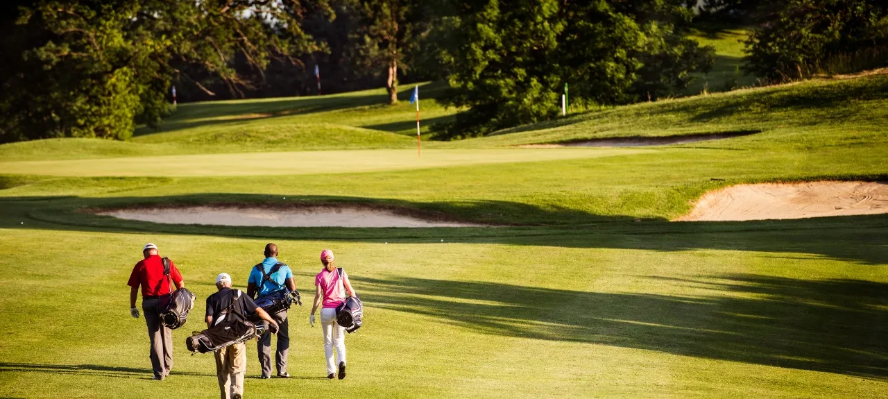 Four golfers carrying their bags down a green fairway towards a putting green with a flagstick