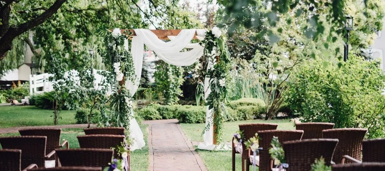 Outdoor garden wedding ceremony with a timber arch draped in white fabric and rows of chairs