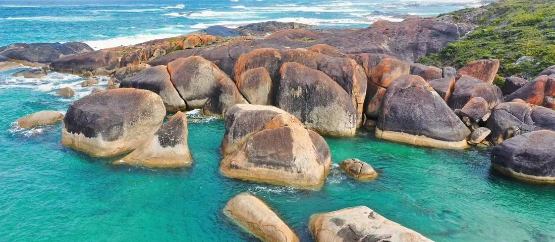 An aerial view of large smooth granite boulders grouped in clear turquoise water along a rocky south coast shoreline