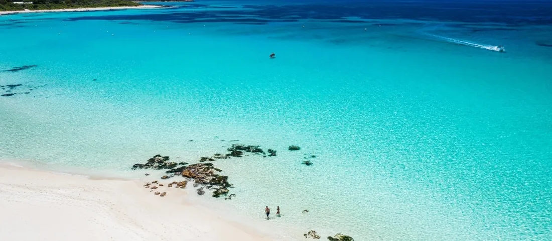 An aerial view of a wide white-sand beach meeting crystalline turquoise shallows where two people wade near the shore