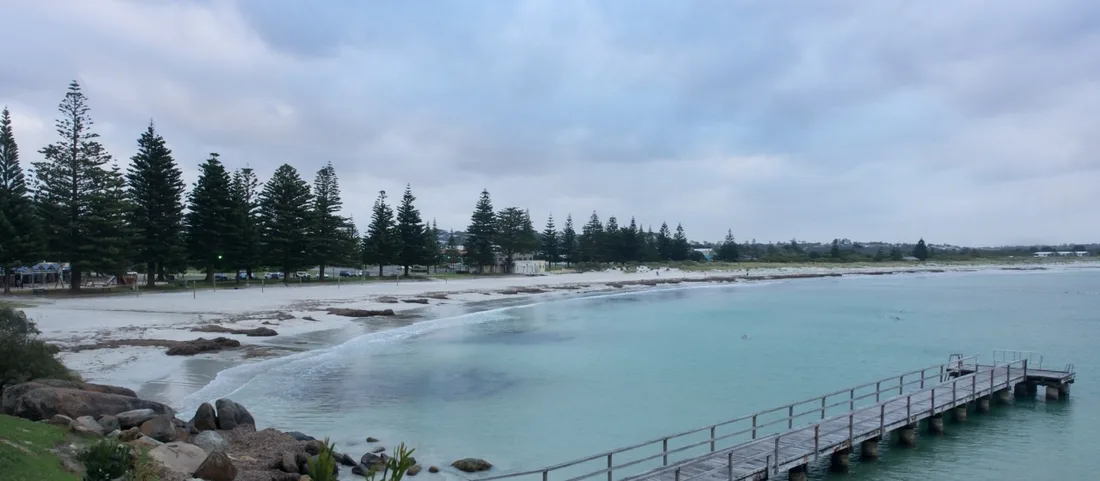 A calm bay with pale sand, a long timber jetty and Norfolk Island pines lining the foreshore under an overcast sky